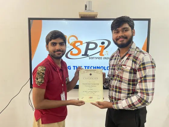 A trainer handing a certificate to a student in front of the Softpro India screen