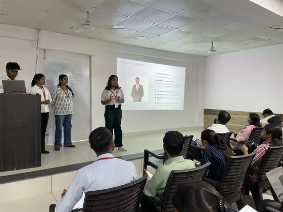 A team presenting to a seated classroom beside a slide introducing Softpro's founder director