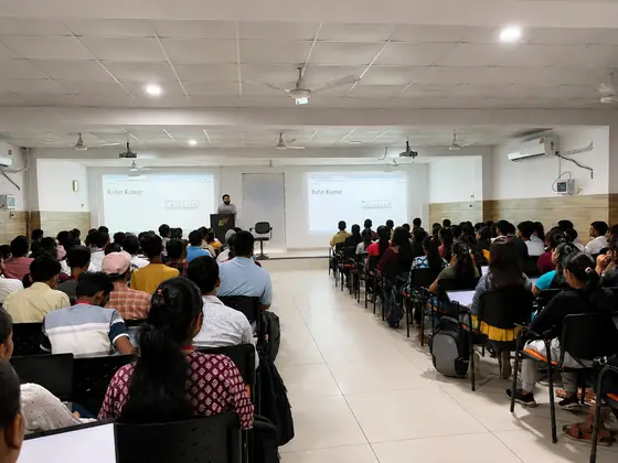 A lecture hall seen from the back, two projector screens lit and a trainer at the lectern
