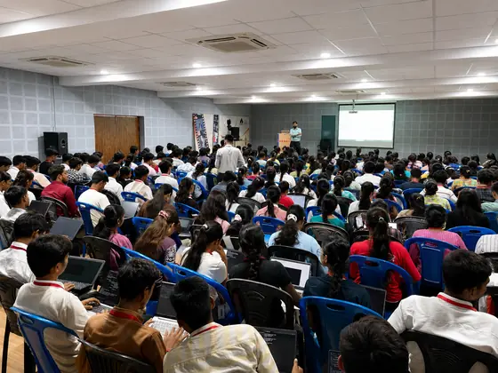 A hall of students photographed from the back rows, the projector screen lit at the far end