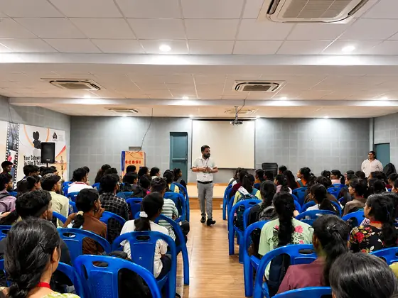 A trainer at the front of a college auditorium speaking to a seated batch