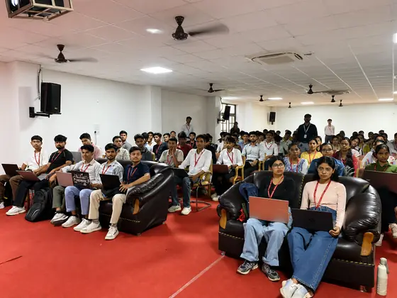 A red-carpeted auditorium with students on the front sofas and rows filling the room behind
